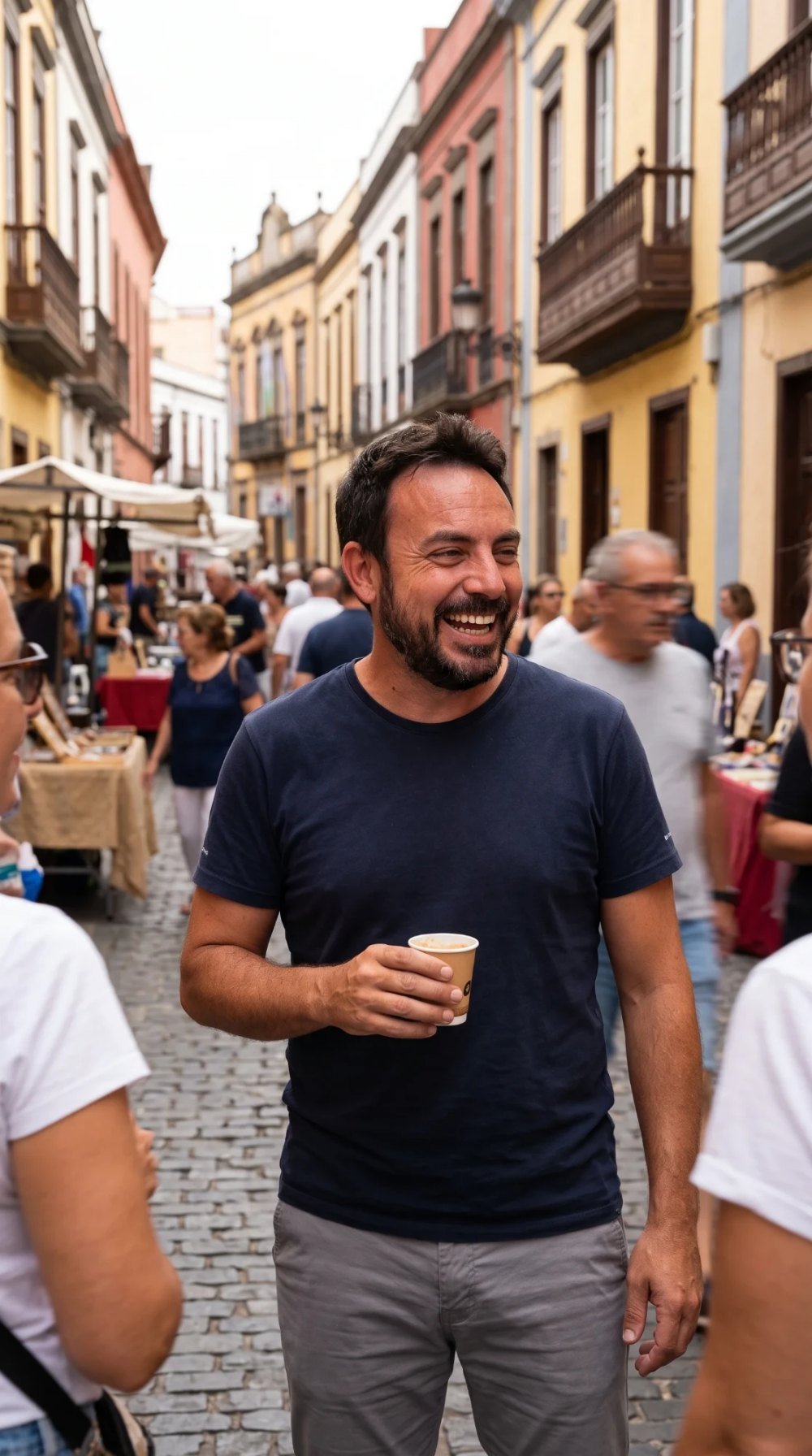Roberto Mickel, riendo con un café en la mano, en una calle empedrada con balcones canarios y puestos de mercado